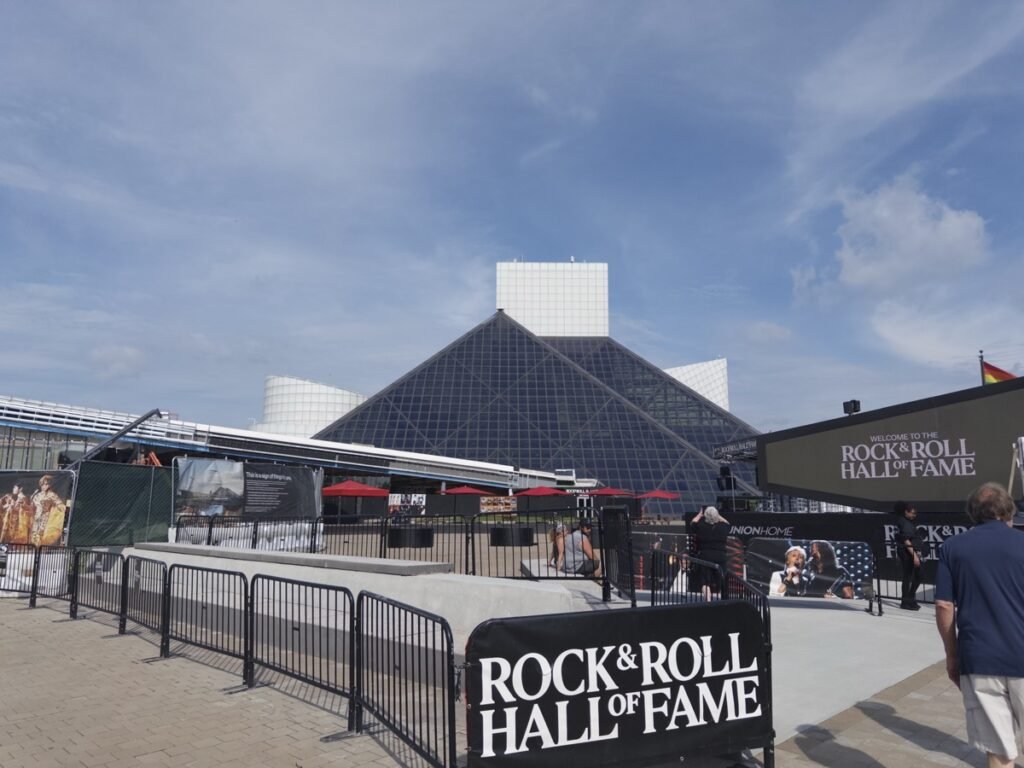 青空の下に建つRock & Roll Hall of Fameのガラス張りのピラミッド型の建物。手前に館名を記した黒い看板と、赤いパラソルが並ぶ屋外テラス