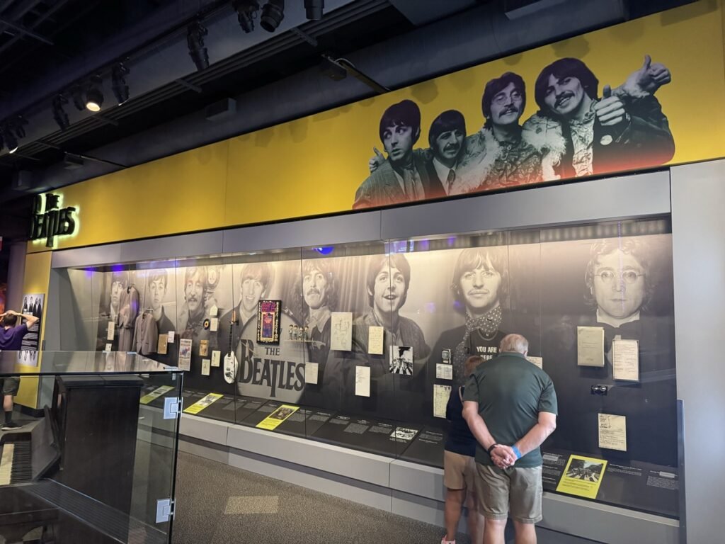 ザ・ビートルズの展示コーナー。黄色い壁に4人の等身大パネルが立ち、ガラスケースにメンバーの白黒写真や直筆資料が並び、来館者2人が見入っている