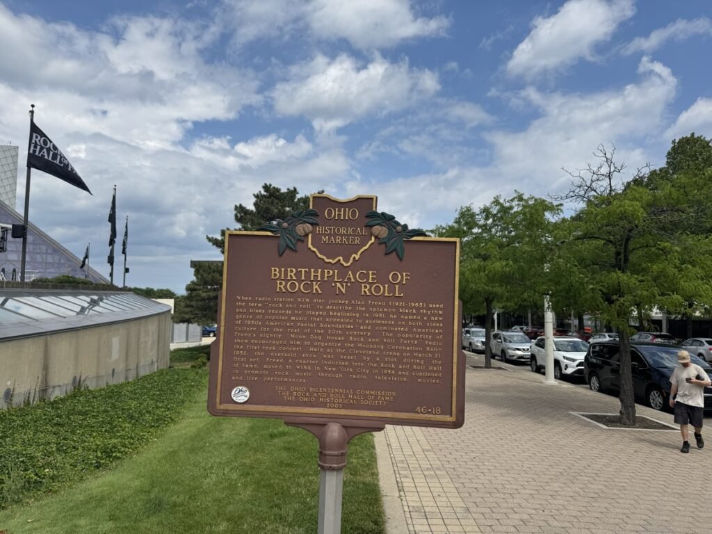 「BIRTHPLACE OF ROCK 'N' ROLL」と題されたオハイオ州の歴史案内板。ラジオDJがロックンロールという言葉を広めた経緯が英文で記され、背後にRock Hallの旗と建物が見える