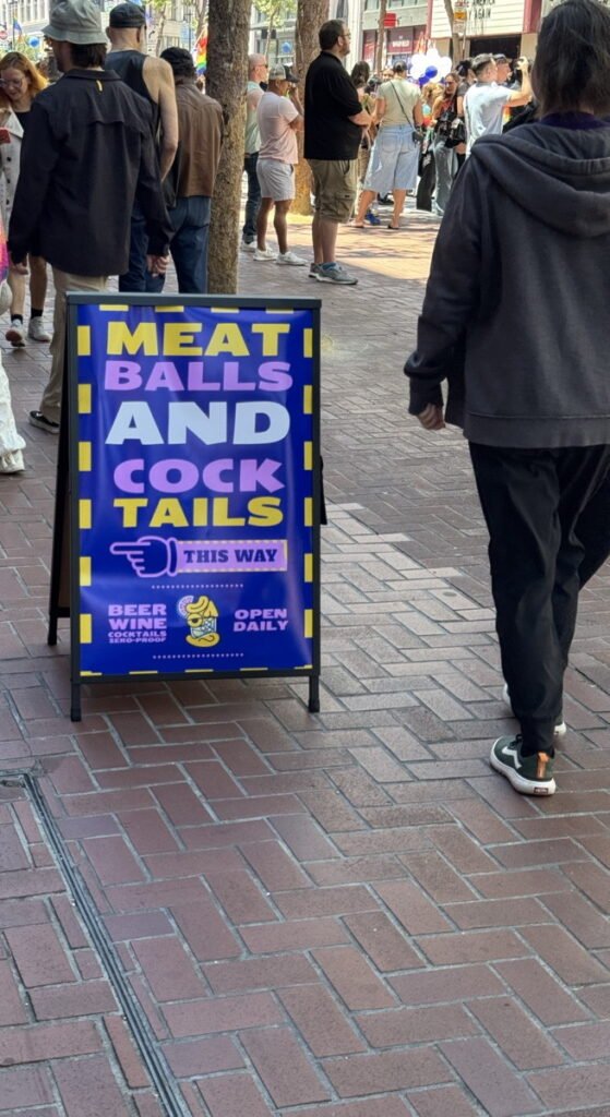 レンガ敷きの歩道に立てられたA型看板。青地に黄色い枠で「MEAT BALLS AND COCK TAILS」の文字が5行に分けて大きく書かれ、指差しマークと「THIS WAY」、下部に「BEER WINE COCKTAILS」「OPEN DAILY」の表記がある。左端は少し切れているが全体は読み取れる