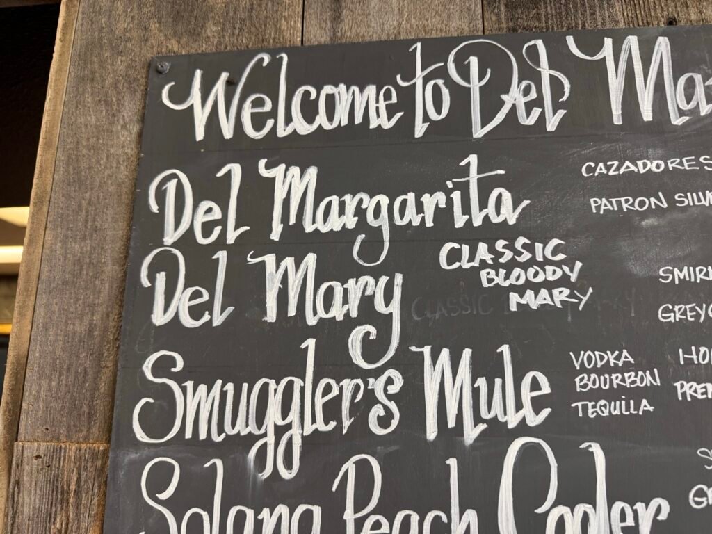 木枠の黒板に白い手書き文字で書かれた場内バーのドリンクメニュー。「Del Margarita」「Del Mary」「Smuggler's Mule」などの名前が並ぶ