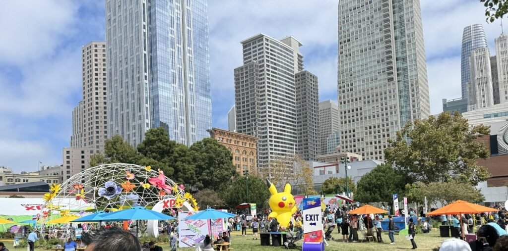 無料エリア「Pokémon XP」の芝生広場。巨大な黄色いピカチュウのバルーンやカラフルな傘、屋台やピクニックテーブルに集まる来場者、背景にサンフランシスコの高層ビル群と青空