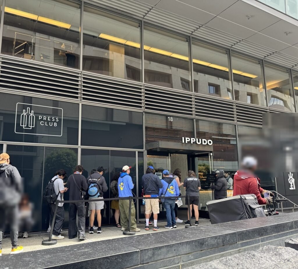 ビル1階の一風堂（IPPUDO）入口前の歩道にできた長い行列。グッズを持った来場者風の人が多く、左には「PRESS CLUB」の看板がある