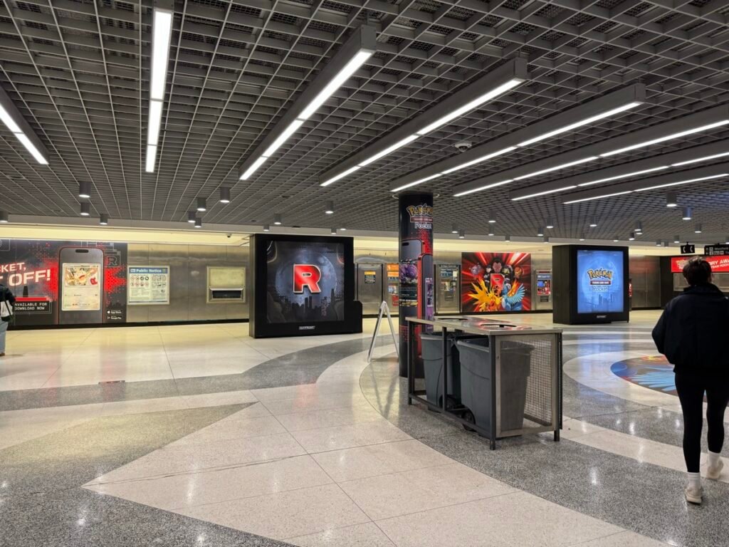 サンフランシスコのPowell駅構内。デジタルサイネージにロケット団の赤い「R」やポケモンカードアプリの広告が表示され、奥を通行人が歩いている