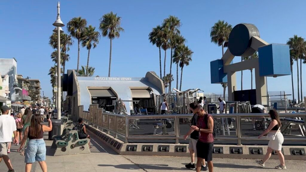 「MUSCLE BEACH VENICE」の看板が立つヴェニスの屋外ジム。青空の下で人々がバーベルやダンベルで筋トレしている