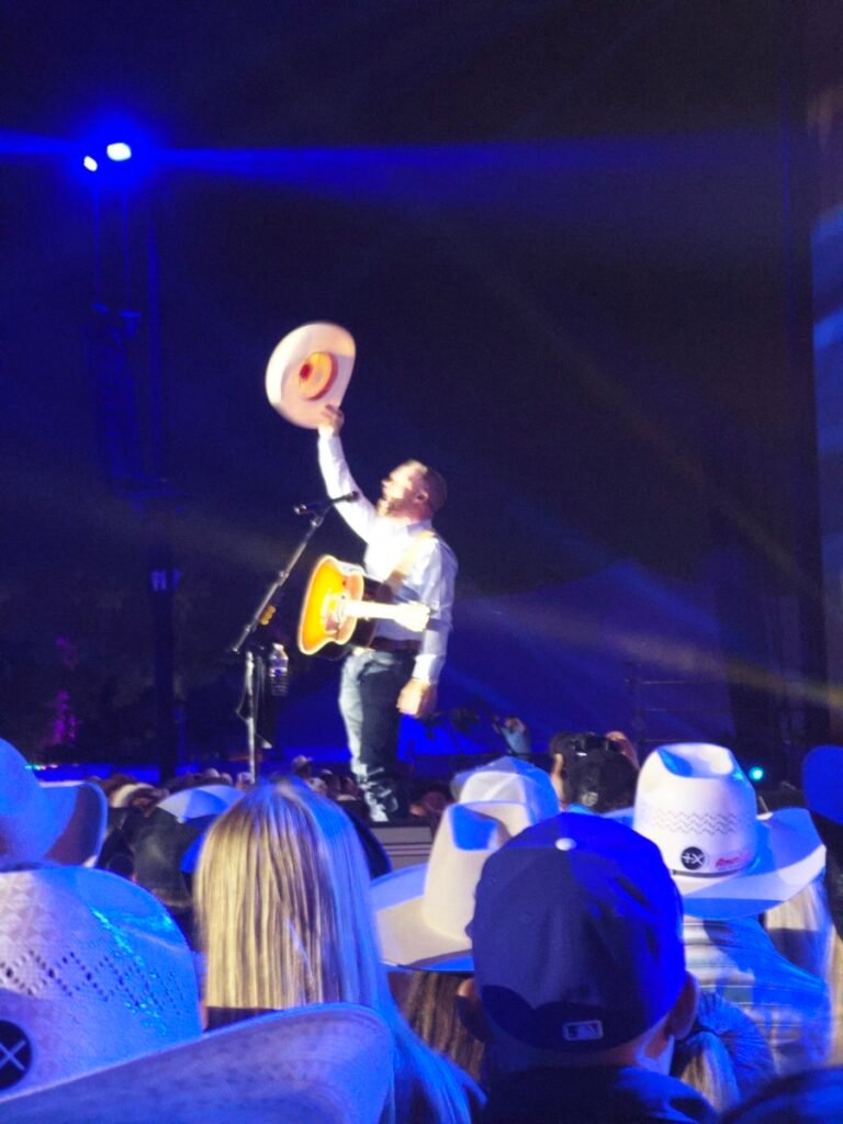 夜のステージで白いカウボーイハットを高く掲げるCody Johnson。手前には後ろ姿の観客のカウボーイハットが並び、青い照明に照らされている