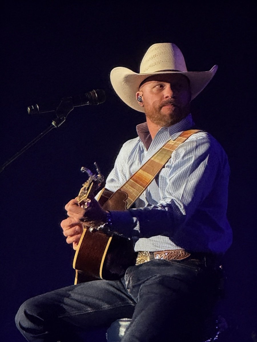 スツールに座り、白いストローハットに青いシャツ姿でアコースティックギターを弾くCody Johnson。マイクスタンドの前、青い照明のステージで歌っている