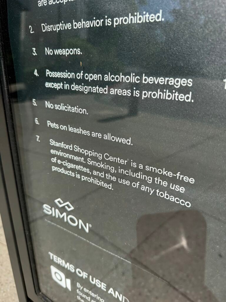 Stanford Shopping Center の黒地の館内ルール掲示を斜めから撮った写真。項目7に「smoke-free environment」とあり、電子タバコを含む喫煙とタバコ製品の使用が敷地内で禁止(prohibited)と英語で書かれている