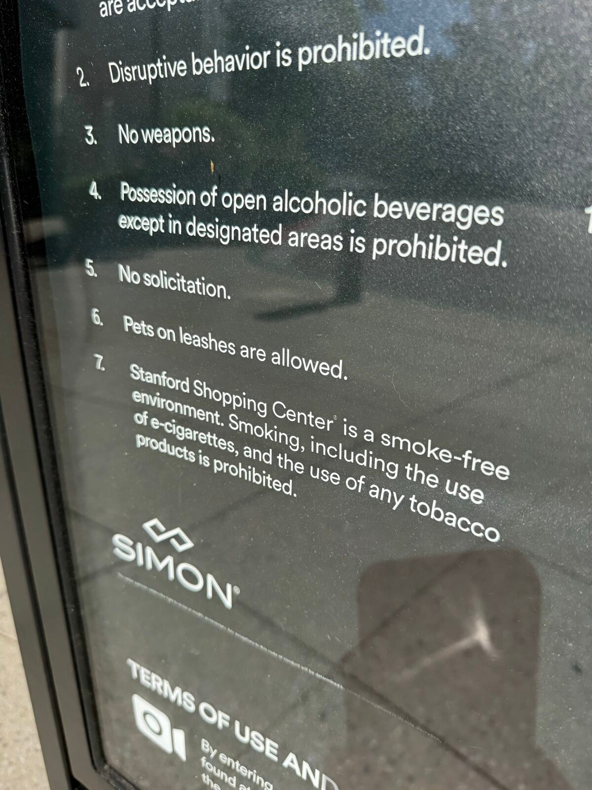 Stanford Shopping Center の黒地の館内ルール掲示を斜めから撮った写真。項目7に「smoke-free environment」とあり、電子タバコを含む喫煙とタバコ製品の使用が敷地内で禁止（prohibited）と英語で書かれている