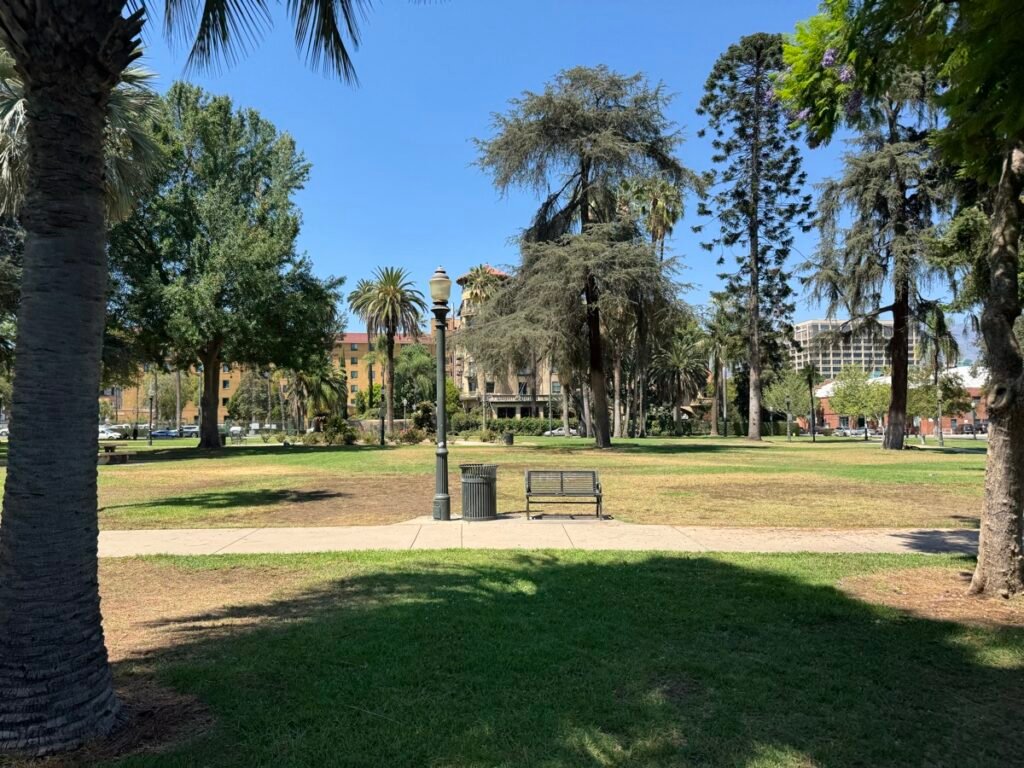 晴れた夏の日のPasadenaのCentral Park。芝生の遊歩道沿いに街灯とベンチがあり、ヤシや木々の奥にCastle Greenの建物が見える