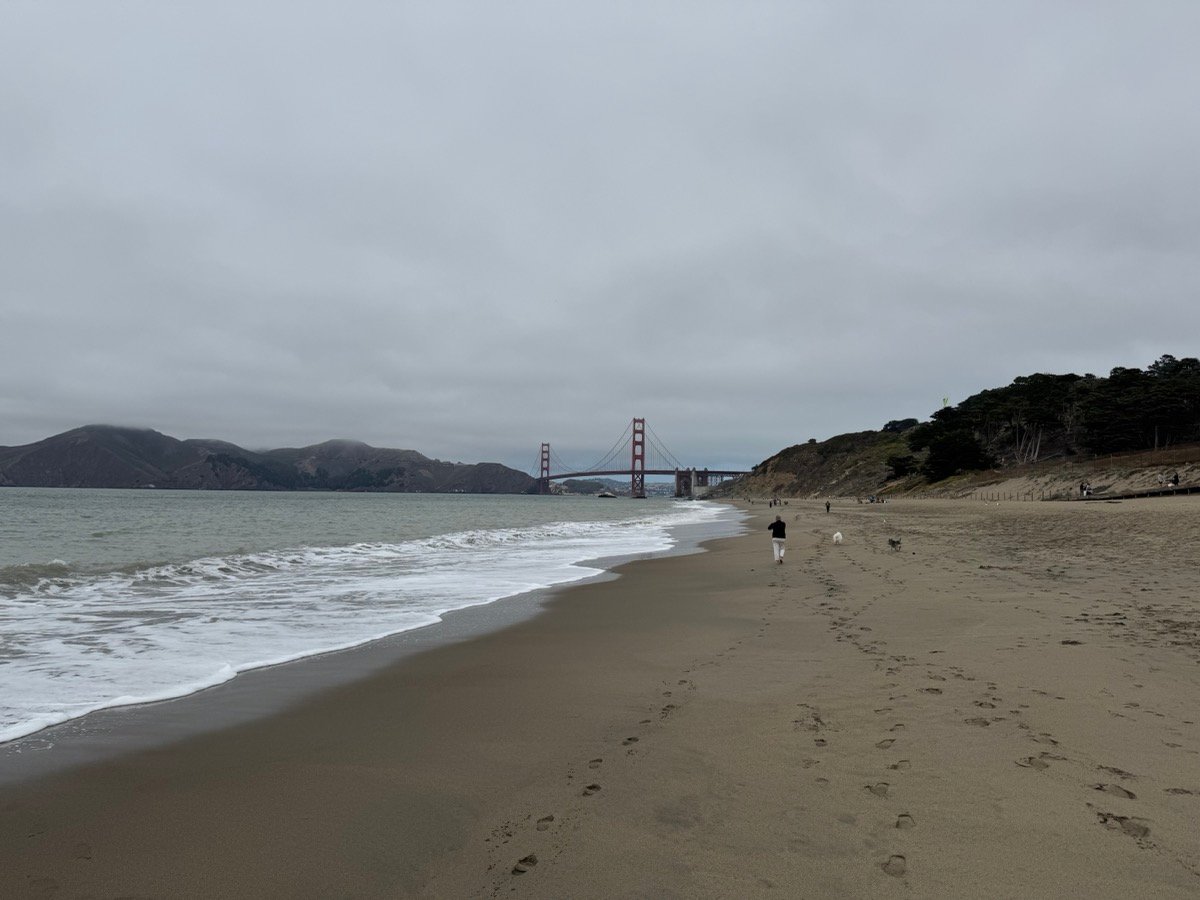 Baker Beachの砂浜越しに見えるゴールデンゲートブリッジ。波打ち際を歩く人と足跡が続く曇り空の景色
