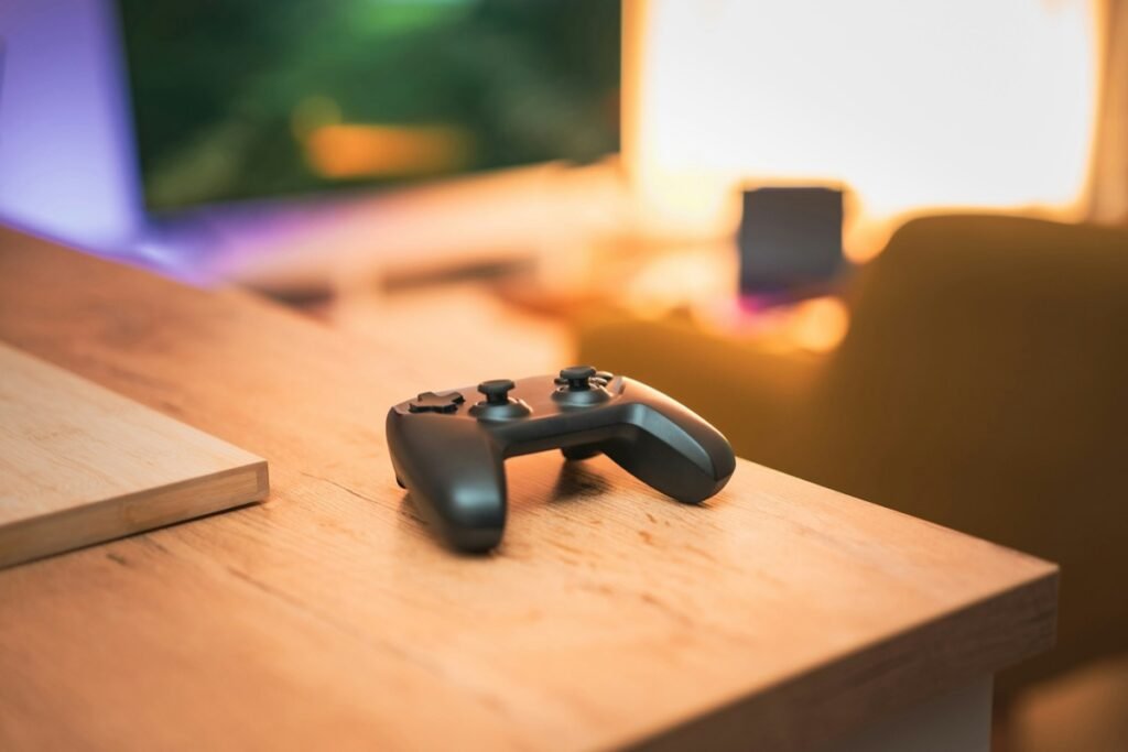 木目のテーブルの角に置かれた黒いゲームコントローラー。奥にはぼやけたモニターと暖色の照明が写っている