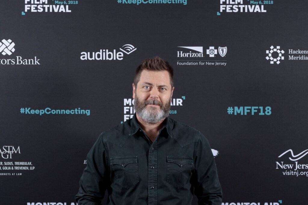 ニック・オファーマンが白髪交じりの顎ひげ姿で、Montclair Film Festivalのバックドロップの前に立っている