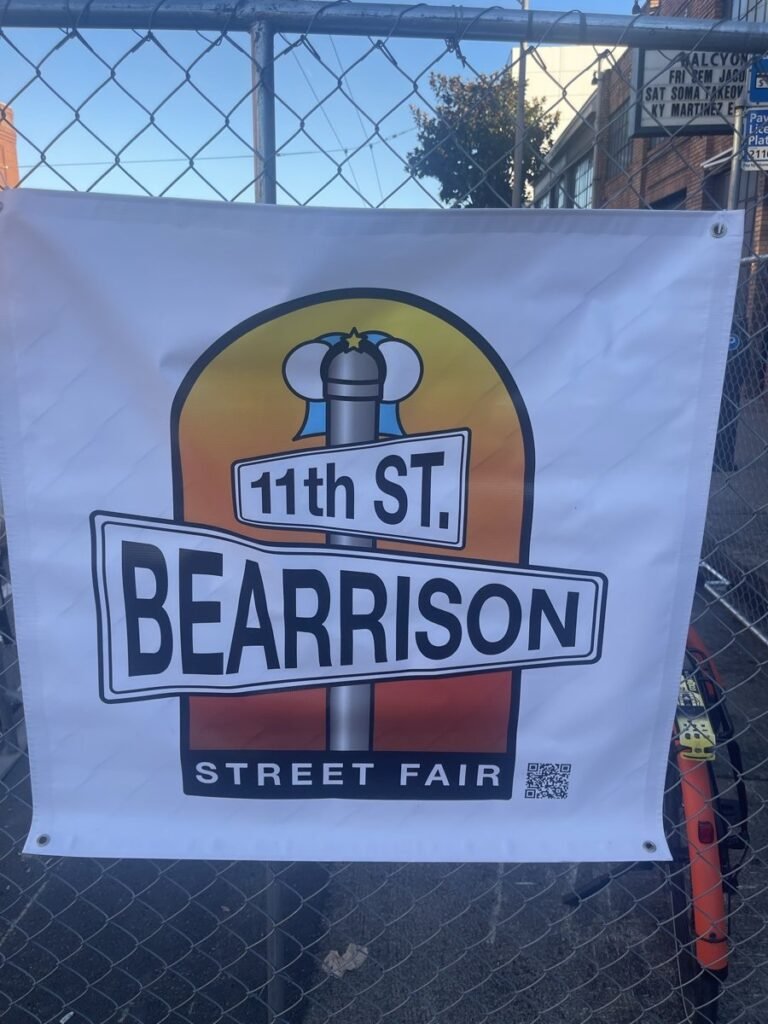 金網のフェンスにかけられたベアリソン・ストリートフェアのバナー。「11th ST.」と「BEARRISON」の道路標識を組み合わせたロゴの下に「STREET FAIR」の文字が入っている