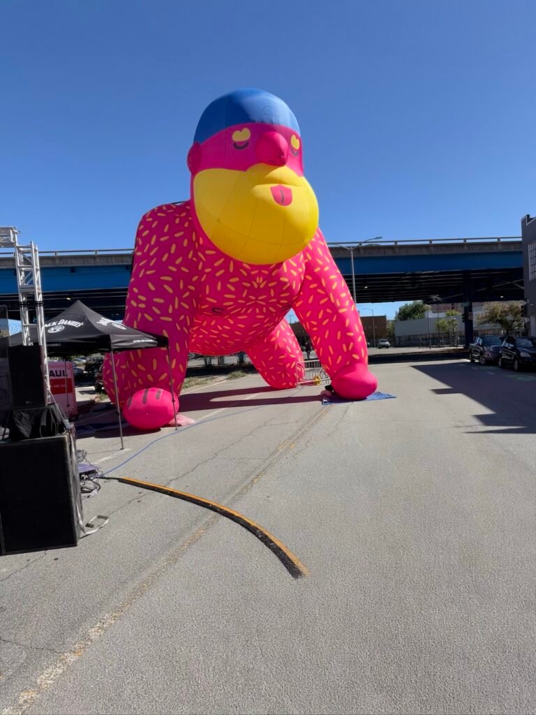青空の下、高架道路の手前の通りに置かれたピンクの巨大なクマのバルーン。青い帽子に黄色いヒゲ、舌を出した顔で四つんばいになっていて、手前にはスピーカーとテントが見える