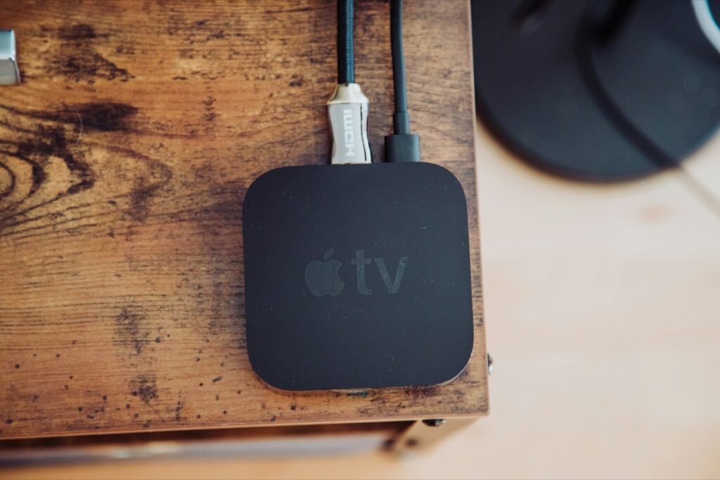 木目のテーブルの上で、HDMIケーブルと電源ケーブルにつながれた黒いApple TV本体を真上から写したイメージ写真(新型ではない旧世代のモデル)