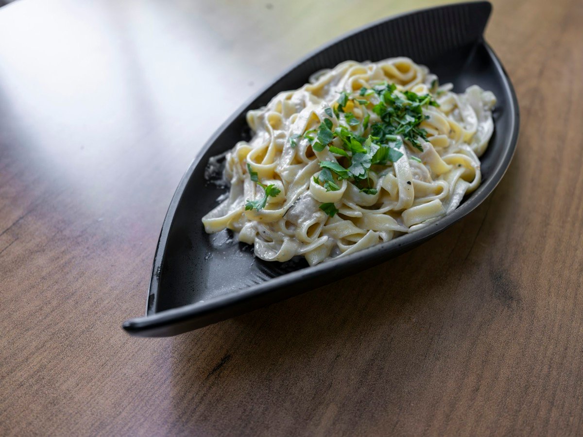 レストランで頼むパスタのイメージ（素材写真）。木目のテーブルの上、葉っぱの形をした黒いお皿に、刻んだパセリをのせたクリームソースのフェットチーネが盛られている