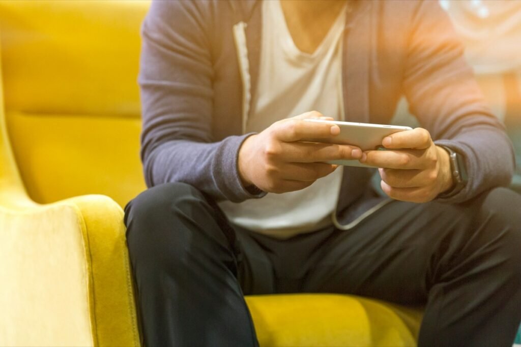 スマホ派のイメージ(素材写真)。黄色いソファに座り、横向きにしたスマホを両手で持って動画を見ているような手元