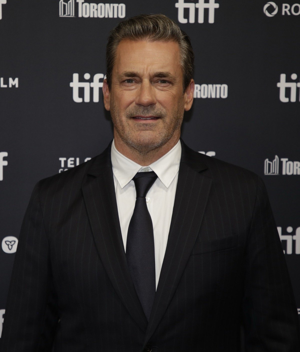 白髪まじりの髪をなでつけ、無精ひげのジョン・ハムが、ストライプのダークスーツに白シャツと黒ネクタイ姿で、トロント国際映画祭(TIFF)のロゴが入ったバックドロップの前に立ち、軽くほほえんでいるポートレート